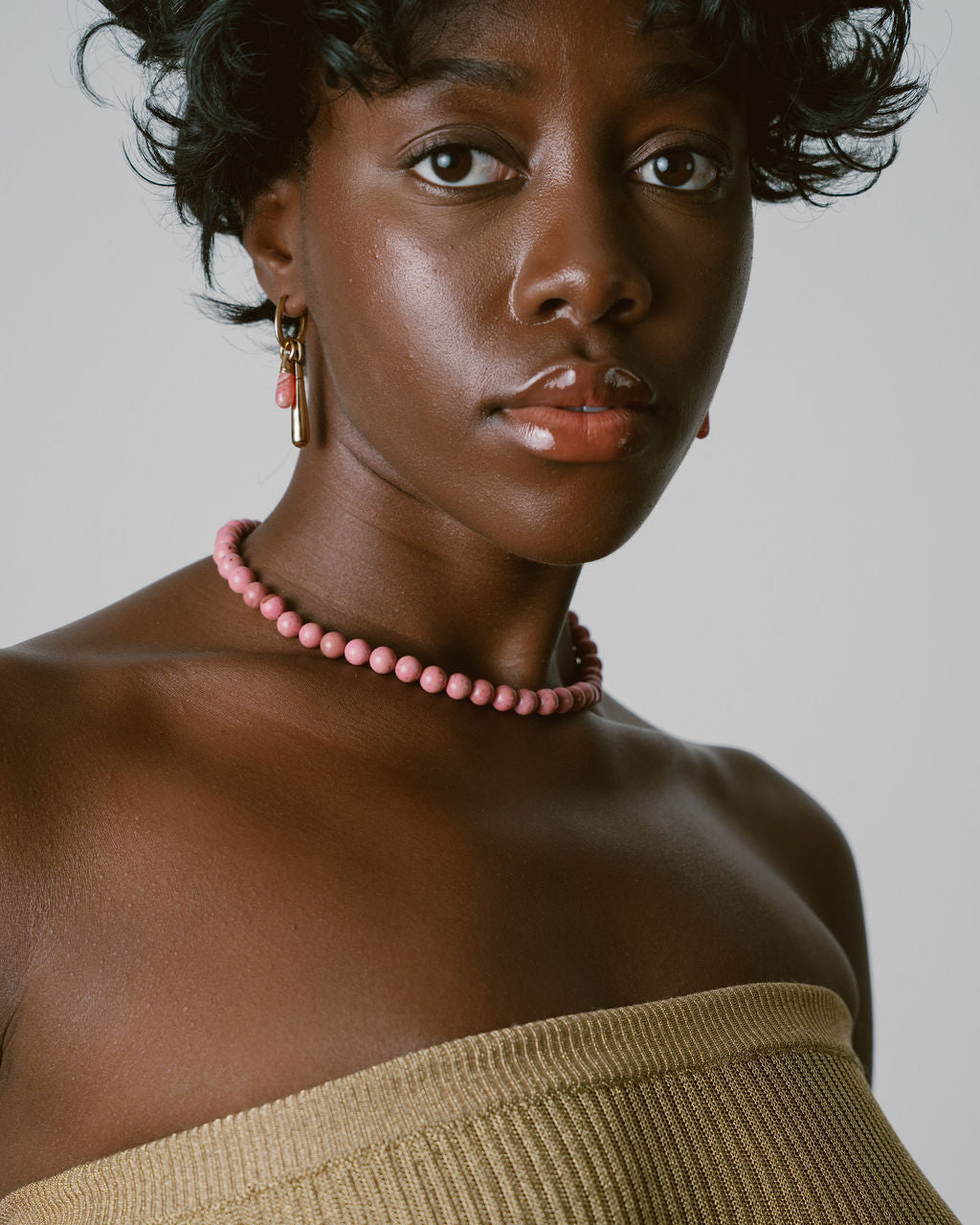 A close-up portrait of a model wearing a pink beaded choker necklace and matching drop earrings, paired with a beige ribbed strap top against a neutral gray background.