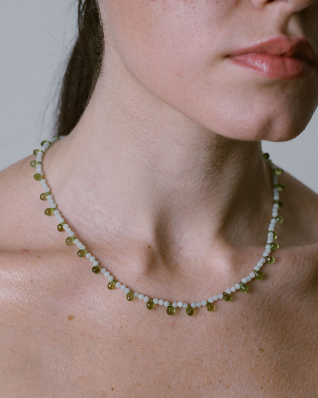 A close-up of a model's neck and upper chest wearing a delicate beaded necklace with small white beads and olive green accents.
